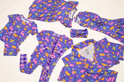 A coordinated baby clothing set laid flat on a light cream background, including a purple long-sleeve romper, matching long-sleeve top and pants, a matching headband, and a zip-front footless sleeper. The fabric has an allover mermaid print in pink, orange, yellow, and blue, with some plaid trim details.
