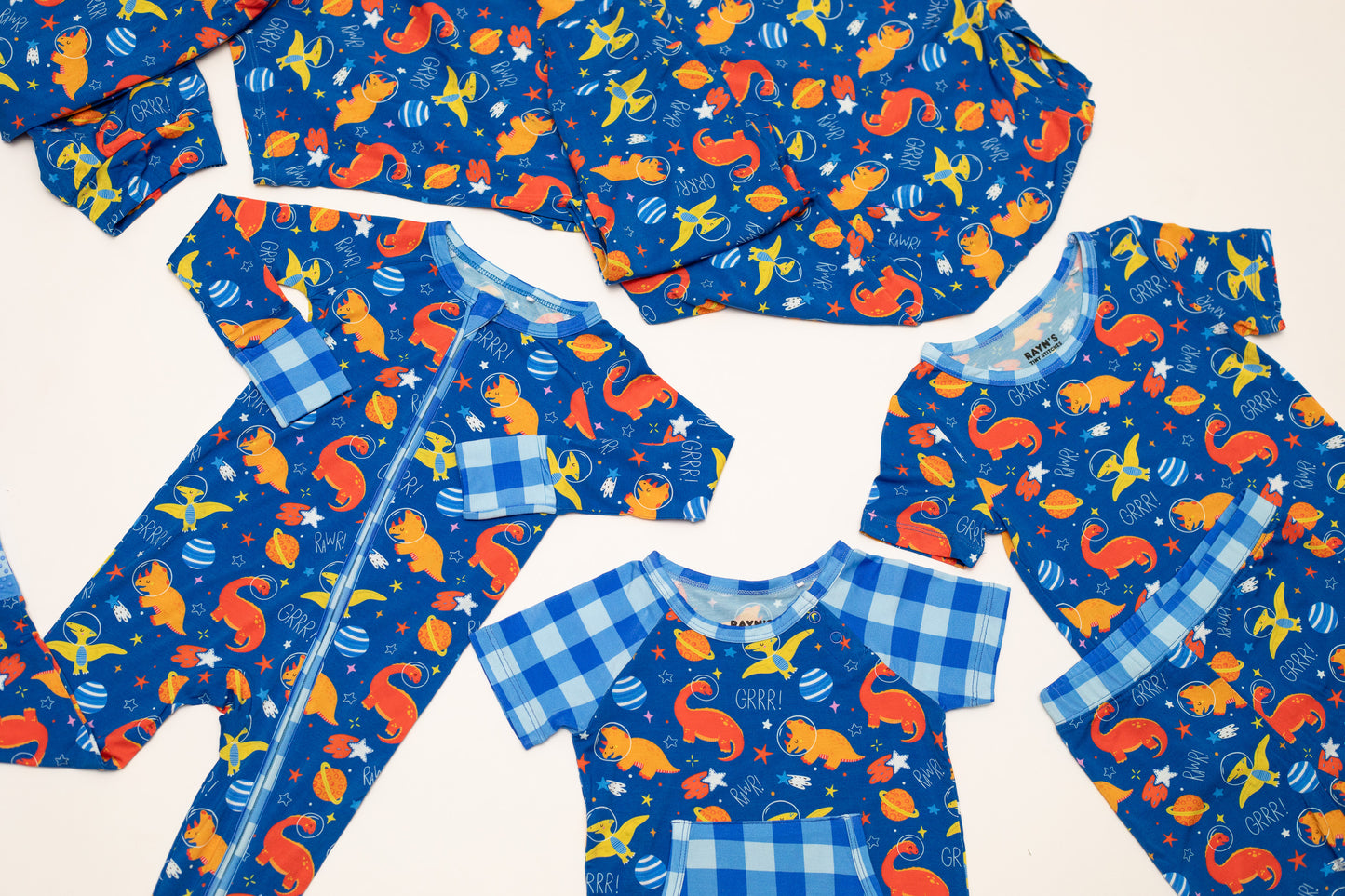 A coordinated baby clothing set laid flat on a light background, including a long-sleeve zip bodysuit and several short-sleeve tops. The fabric has a blue base with a playful print of orange squirrels, yellow birds, blue fish, and white stars, plus light blue gingham trim.