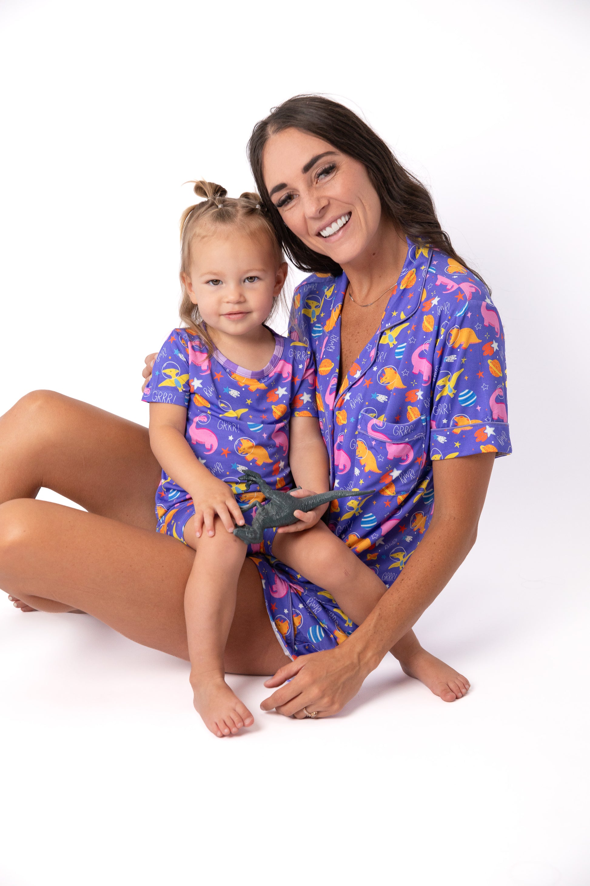A woman and a young girl sitting together on a white studio background, both wearing matching short-sleeve pajama shirts and shorts with a colorful dinosaur print on a purple base.