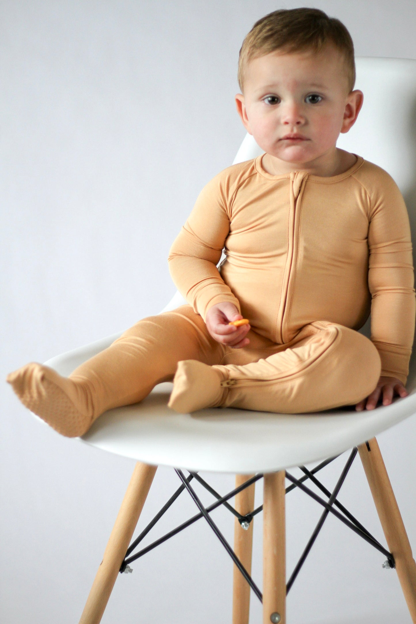 Child wearing a beige onesie sitting on a white chair with a light gray background